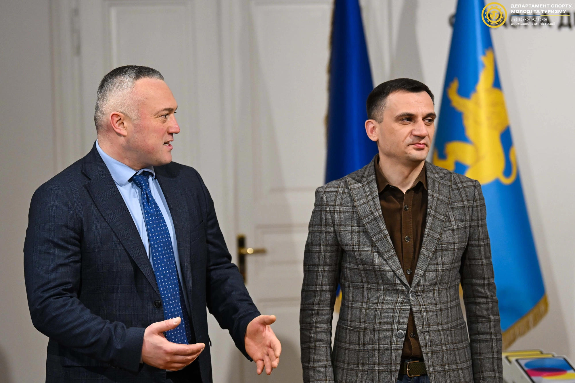 Двоє чоловіків у ділових костюмах стоять на тлі прапорів України в офіційній обстановці