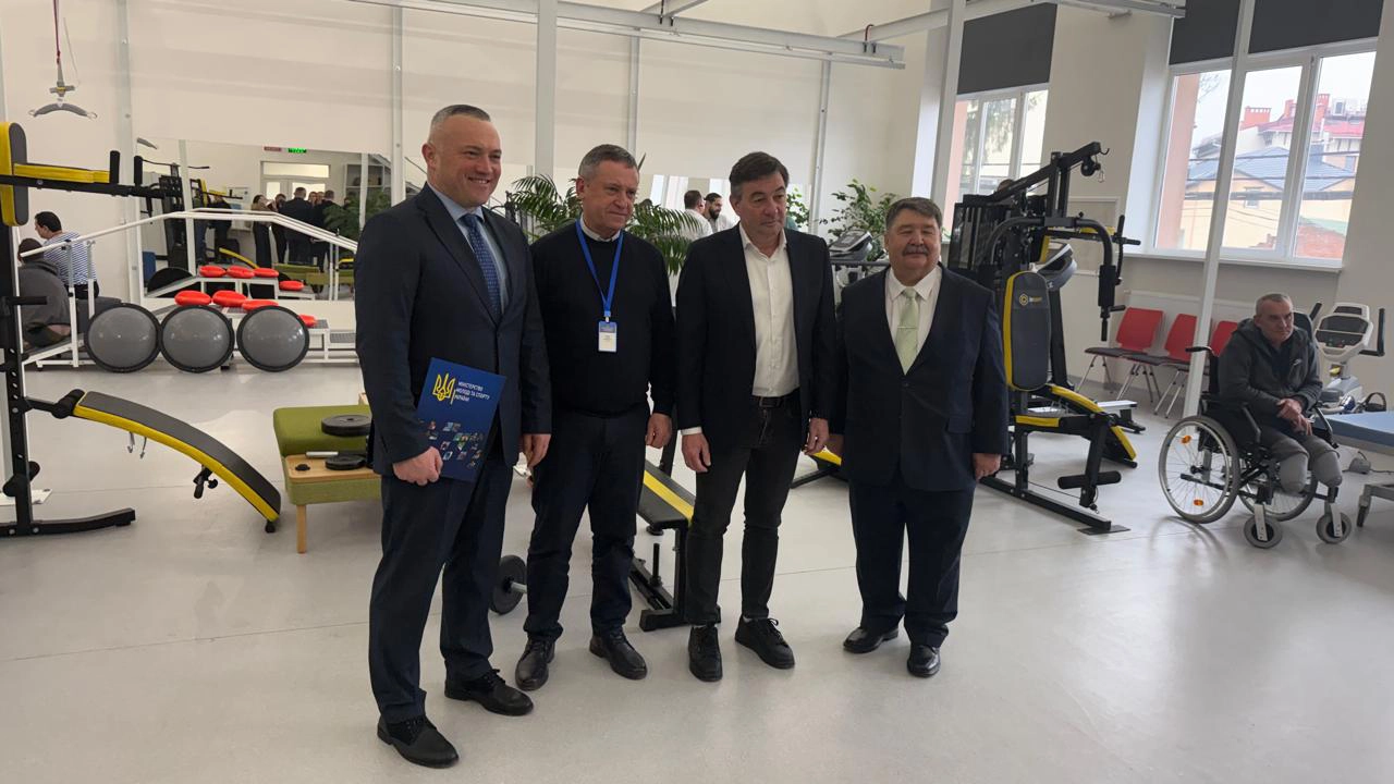 Чотири чоловіки стоять у спортивному залі, поруч тренажери та людина в інвалідному візку