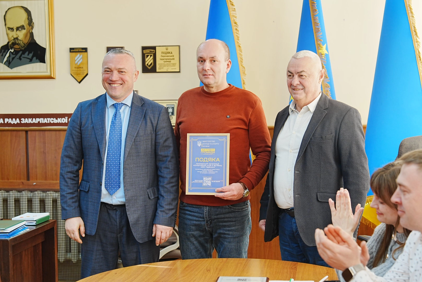 Три чоловіки в ділових костюмах стоять разом, один тримає подяку, на задньому плані прапори України