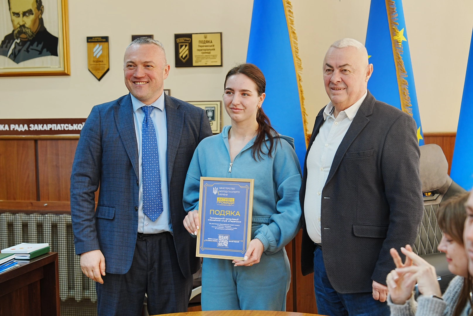 Три людини на церемонії нагородження, жінка тримає подяку, позаду прапори України