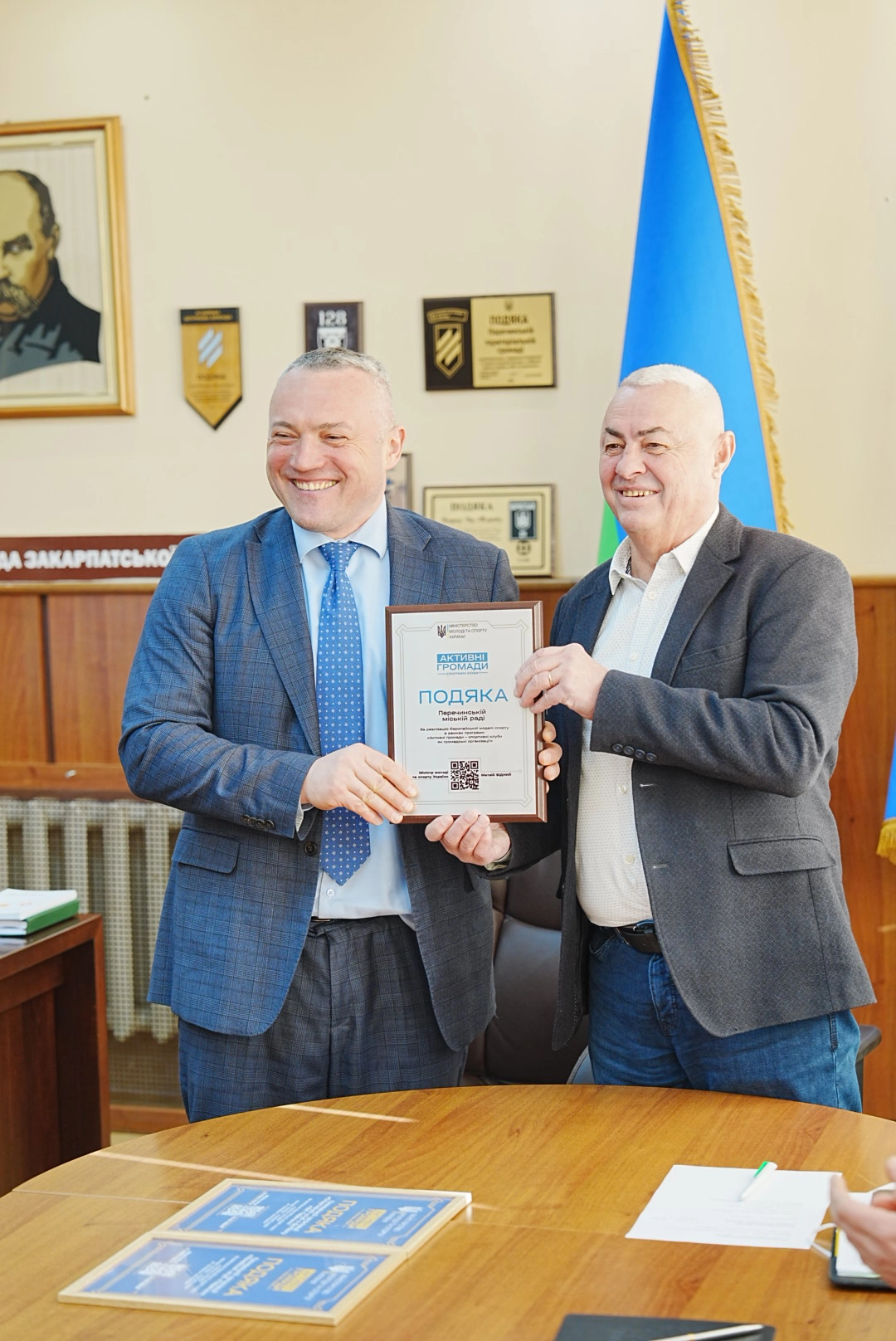 Двоє чоловіків у ділових костюмах тримають подяку в офісі на тлі прапора України